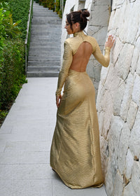 Wearing an Aurora Gold Maxi Dress, a woman exudes effortless glamour as she stands on a stone pathway facing a stairway. The backless design of her long-sleeved gown accentuates her elegant silhouette, with green foliage bordering the path to infuse the scene with natural beauty.