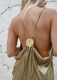 The Elara Gold Maxi Dress features a shimmering gold fabric with thin straps and a playful low-back design, accented by a round gold decorative piece. Photographed against a textured white wall, this metallic masterpiece embodies elegance with its open back detail.