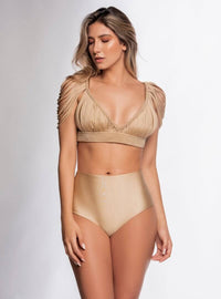A woman with long blonde hair is wearing a minimalist beige two-piece outfit. The top showcases textured draped sleeves and a deep V-neckline, which she has paired seamlessly with the Selene High Waisted Bikini Bottom. She stands against a neutral background, gazing slightly to her right.