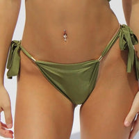 Sunlight illuminates a person wearing the Hydra Bikini Bottom, showcasing its adjustable side ties. The scene centers on the lower torso, accentuating a belly button piercing adorned with a delicate charm. The setting is enveloped in bright, neutral tones, embodying an essence of Italian craftsmanship.