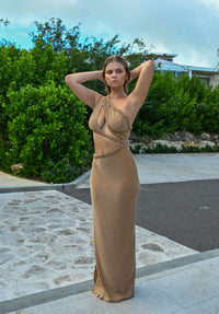 Amidst a backdrop of lush greenery and a cloudy sky, a woman stands self-assuredly on a stone path. She wears the striking Tami Gold Maxi Dress, characterized by its tan color, asymmetrical cutouts, and detailed braided accents. With her hands gently raised to her head, she exudes confidence outdoors.