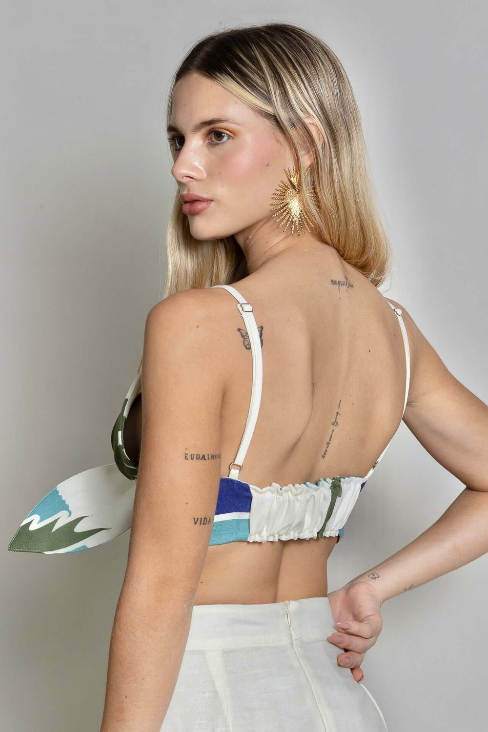 A person with long blonde hair, wearing the colorful Panama Top & Maxi Skirt, exudes tropical vibes as they look over their shoulder. Adorned with multiple tattoos and a large star-shaped earring, they stand against a plain light background.