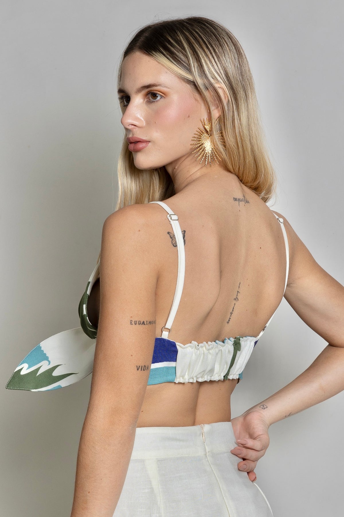 A person with long blonde hair, wearing the colorful Panama Top & Maxi Skirt, exudes tropical vibes as they look over their shoulder. Adorned with multiple tattoos and a large star-shaped earring, they stand against a plain light background.