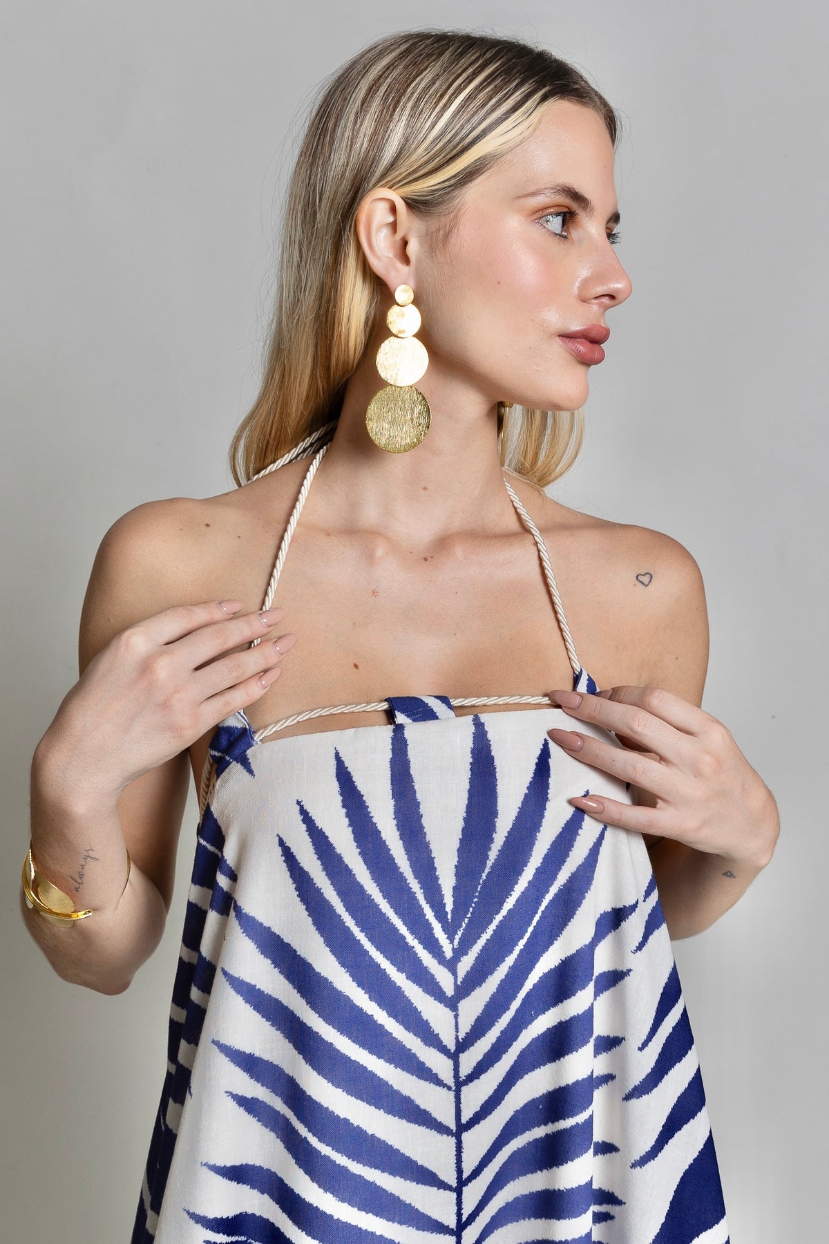 A woman poses in the Rachel Maxi Dress, a stunning blue and white maxi dress featuring a bold leaf pattern. She accessorizes with large gold earrings and a bracelet. Her blonde hair cascades down, highlighting the small heart tattoo on her shoulder. This summer showstopper look makes a statement against the plain background.