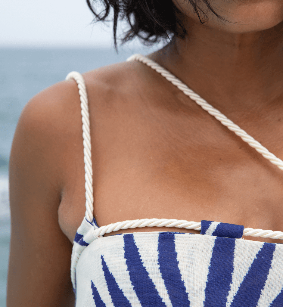 Close-up of a person wearing the Rachel Maxi Dress, featuring a blue and white sunburst pattern with rope-like straps. This summer showstopper stands out against the blurred view of the ocean and sky.