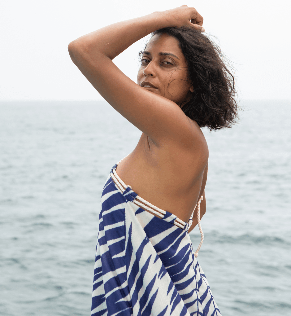 By the ocean, a person with short hair stands wearing the Rachel Maxi Dress—a vibrant halter design that perfectly encapsulates the essence of a summer showstopper. Striking a pose with one arm raised, they gaze into the distance, their serene expression complemented by the breathtaking backdrop of sea and sky.