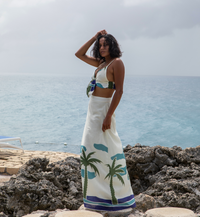 A woman stands on a rocky shoreline, exuding tropical vibes in the Panama Top & Maxi Skirt adorned with palm tree prints. Her versatile outfit complements the ocean backdrop under a cloudy sky.