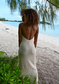 Wearing an Aria Gold Maxi Dress with a plunging neckline, a woman stands on a sandy beach by the turquoise water. Her figure-skimming silhouette is beautifully highlighted against the lush greenery as she faces away, showcasing a tattoo on her upper back beneath the partly cloudy sky.