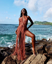 Dressed in the Afrodita Macrame Cover Up With Bikini, an individual stands confidently on rocky terrain by the ocean. The ensemble, featuring brown fringed details reminiscent of Colombian macrame techniques, complements the scene framed by a blue sky and scattered clouds with green hills creating a picturesque backdrop.