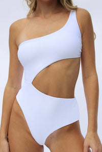 A woman is modeling the Aviva One Piece, an elegant one-shoulder swimsuit crafted from white Italian ribbed fabric, showcasing a fashionable diagonal cutout across the torso. She stands against a plain white backdrop.