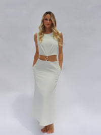 A woman stands barefoot against a white background, wearing the elegant Sorelle Tea Length Dress featuring cut-out sides and a decorative ring at the waist. Her long, wavy blonde hair frames her serene expression as her hands rest in the dress pockets—making it a perfect addition to any resort wardrobe.