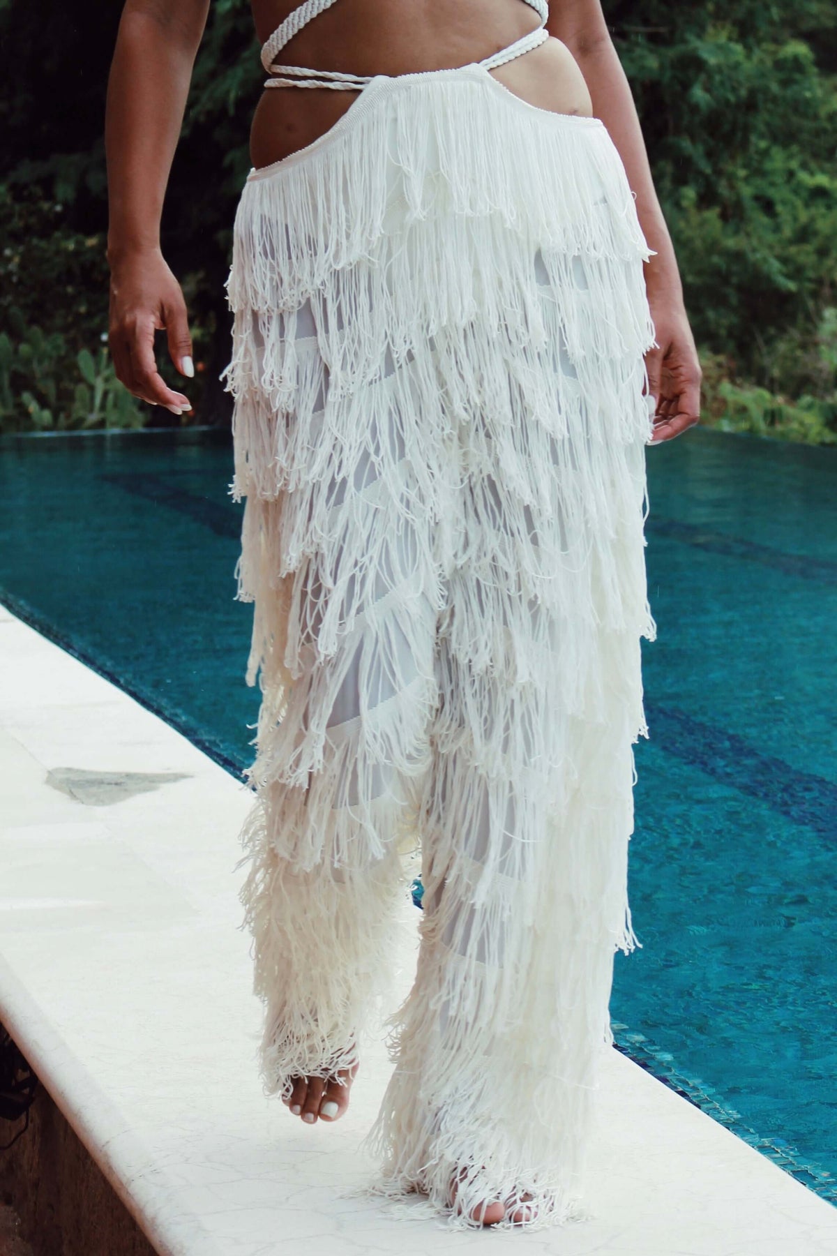 A person strolls beside a pool in the stunning Bali Pants, adorned with fringes and sheer accents. The lush greenery in the background accentuates the elegance of this handcrafted fashion piece.