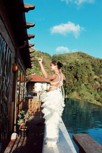 Wearing Bali Pants, a woman in a white fringed outfit, showcasing exquisite Colombian craftsmanship, poses playfully by an infinity pool with lush green hills in the background under a clear blue sky.