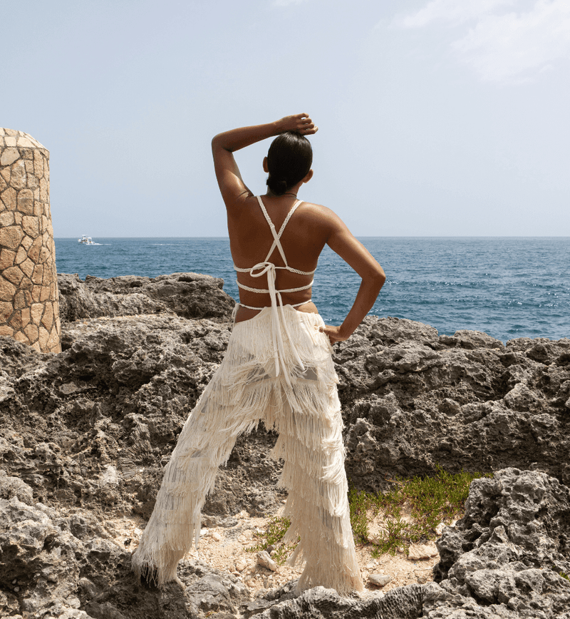 A person stands on rocky terrain facing the ocean, dressed in white Bali Pants with fringed details that exude Colombian craftsmanship. The sky is clear, and the sea stretches to the horizon.