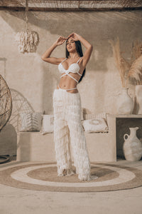 A woman wearing a Bali Handmade Macrame Bikini Top and fringed pants poses in a stylish, earthy-toned room adorned with woven decor and vases. She stands on a circular rug, emanating boho goddess vibes, with a hanging chair and pillows in the background.