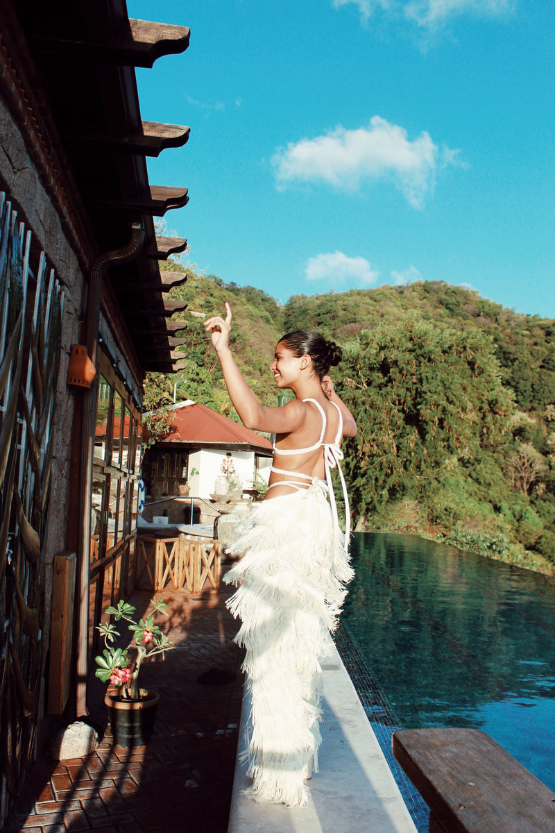 By the pool, amidst lush greenery and under a bright tropical sun, a woman stands in a joyful pose wearing the Bali Handmade Macrame Bikini Top. This white fringed outfit, featuring intricate macrame detailing, perfectly complements the relaxed atmosphere as she looks and points upwards with an exuberant expression.