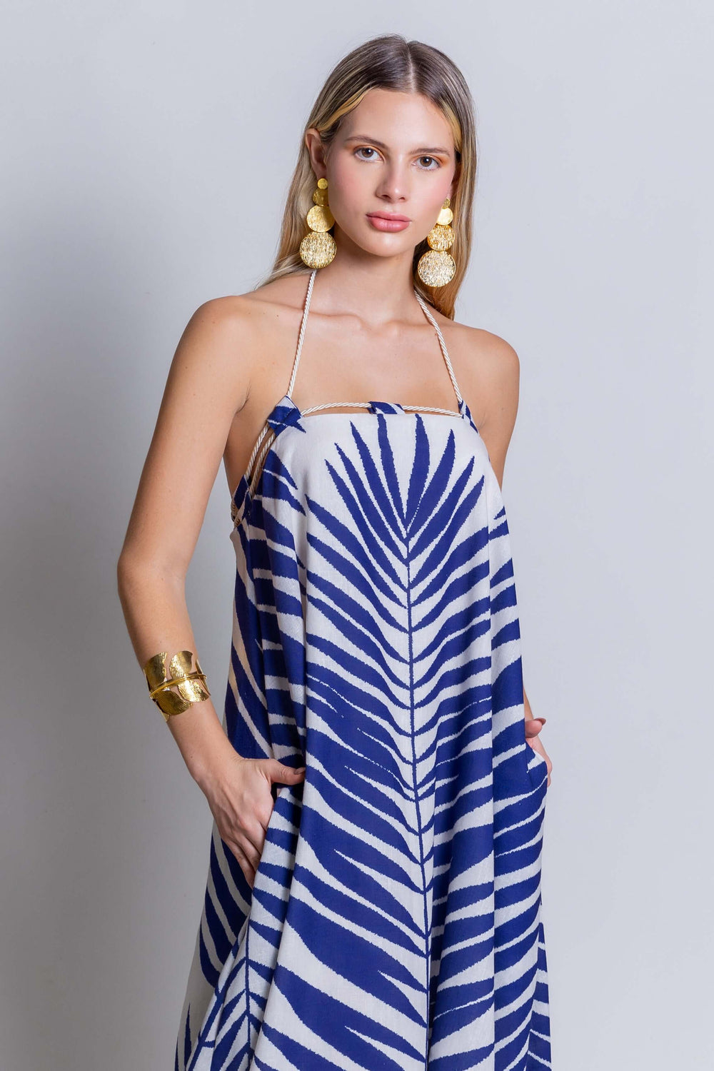 A woman stands against a plain background, wearing the Rachel Maxi Dress, which features a flowing blue and white zebra pattern. She has long hair and is accessorized with large gold earrings and a chunky gold bracelet. Her hands are in the pockets of her vibrant summer showstopper.