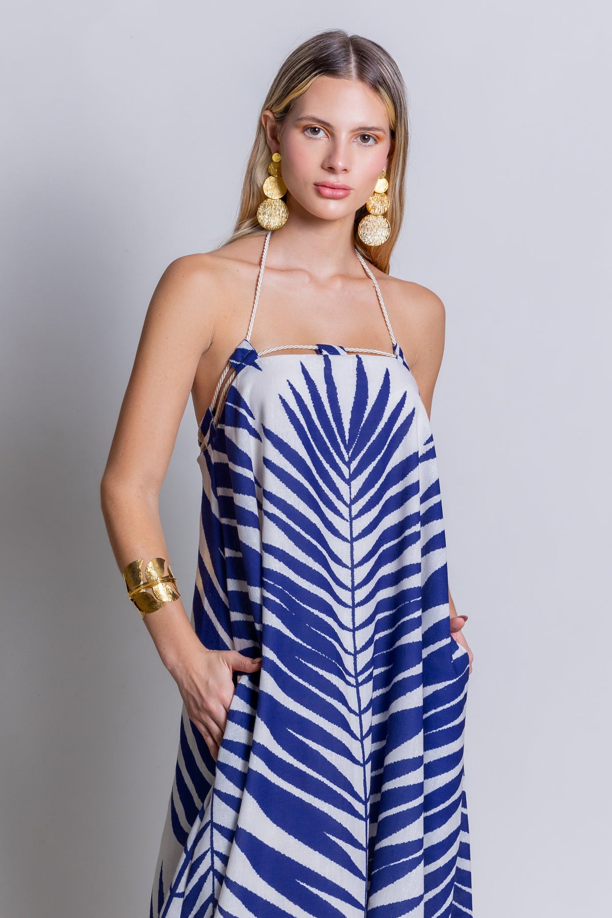 A woman stands against a plain background, wearing the Rachel Maxi Dress, which features a flowing blue and white zebra pattern. She has long hair and is accessorized with large gold earrings and a chunky gold bracelet. Her hands are in the pockets of her vibrant summer showstopper.