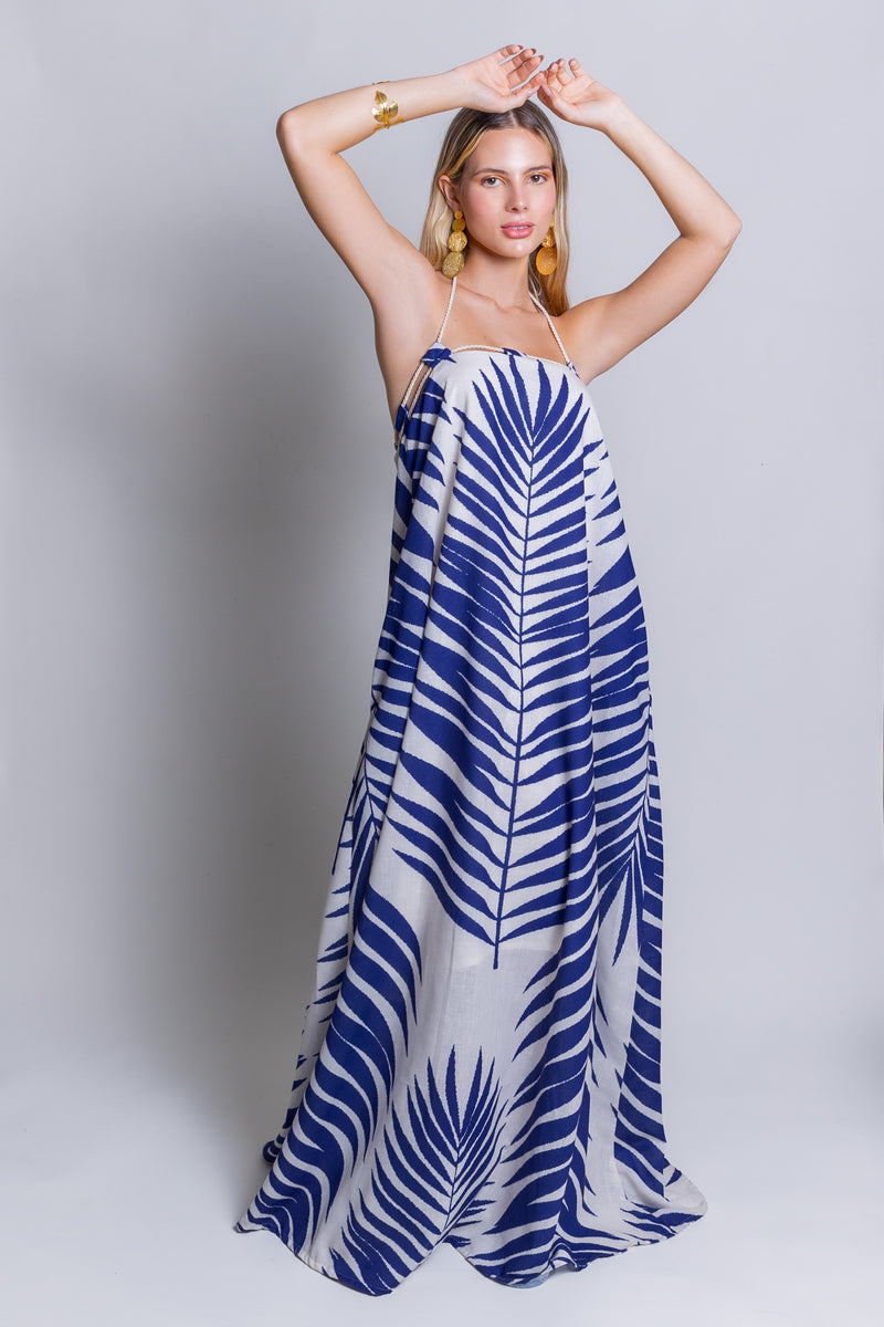 A woman poses with her arms above her head, showcasing the Rachel Maxi Dress adorned in a vibrant leaf pattern. The plain, light grey background highlights her choice of gold earrings and a bracelet, turning this summer showstopper ensemble into an unforgettable look.