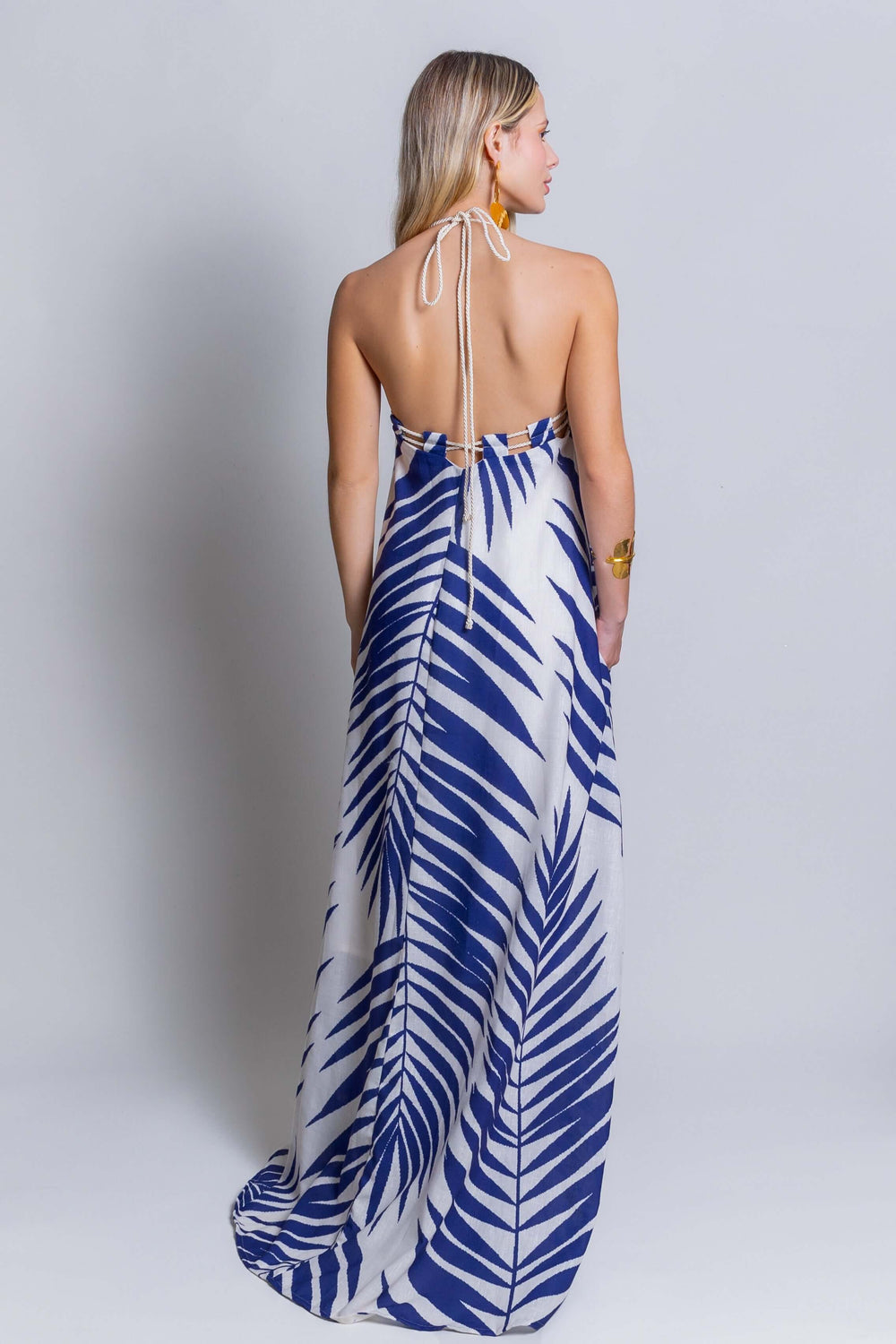 Rear view of a woman with long blonde hair wearing the Rachel Maxi Dress, a stunning backless halterneck dress showcasing a vibrant blue and white palm leaf pattern. She complements her look with a bracelet and earrings, creating a striking summer statement against a plain background.