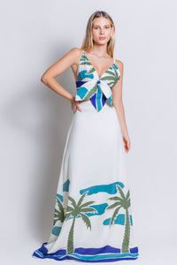 A woman wears the Panama Top & Maxi Skirt, a versatile long white dress set embellished with green palm trees and blue waves. This sleeveless linen ensemble features a tied front detail and a v-neckline, radiating tropical vibes as she stands against a plain background.