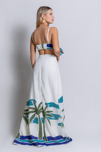 A woman wearing the Panama Top & Maxi Skirt, adorned with tropical palm tree designs, stands sideways against a plain backdrop, exuding tropical vibes with her long skirt and sleeveless top. She accessorizes with large earrings and has long, straight blonde hair.