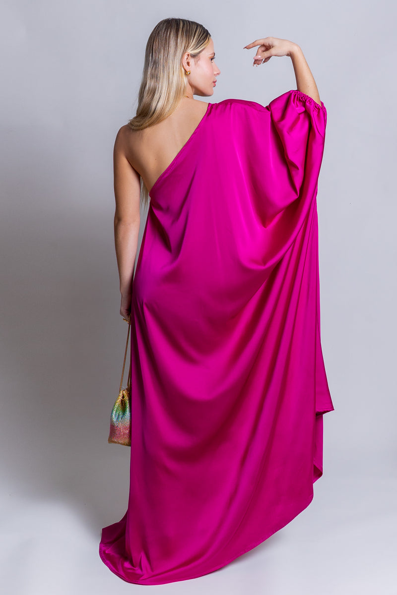 A woman with long blonde hair poses with her back to the camera, wearing a fuchsia pink Capri Maxi Dress featuring a one-shoulder design and a flowing drape. She holds a small colorful handbag and has one arm elegantly raised against a plain light gray background.