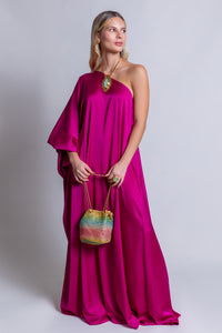 A woman stands against a plain background, wearing the vibrant Capri Maxi Dress in fuchsia pink with a one-shoulder design. She holds a colorful, shimmering drawstring bag and accessorizes with a large pendant necklace and ring, her long, straight hair styled down.