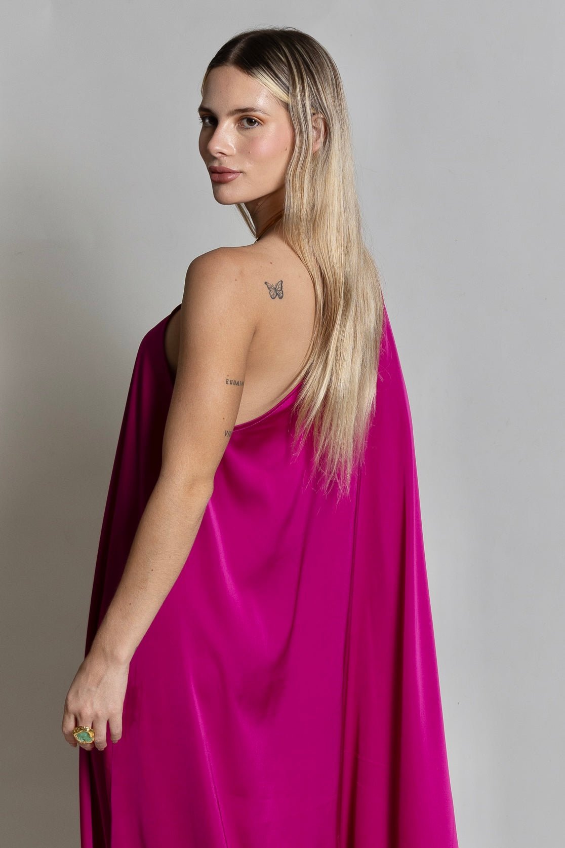 In front of a simple backdrop, an individual with long, blonde hair showcases the striking Capri Maxi Dress in vibrant fuchsia pink. A delicate butterfly tattoo adorns their shoulder as they turn to gaze at the camera.