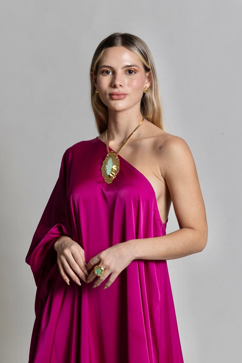 A woman poses confidently in a vibrant fuchsia pink Capri Maxi Dress with an off-shoulder design. She accessorizes with a large pendant necklace and a matching ring, her long, straight hair cascading down as she maintains a serene expression against a simple grey background.