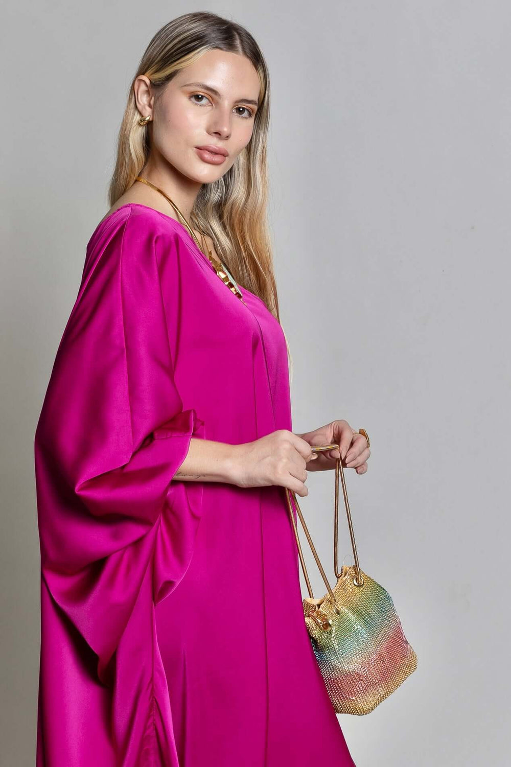 A person with long blonde hair stands against a neutral gray background, wearing the vibrant fuchsia pink Capri Maxi Dress and holding a colorful drawstring bag.