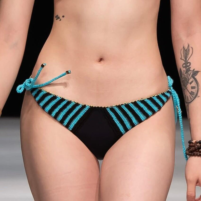 A person wearing the Celeste Crochet Bikini Bottom, adorned with black and blue colors and stylish side ties, struts down the runway, exuding vibes reminiscent of the Amalfi Coast. Their tattoos grace their arm and waist, perfectly complemented by a beaded bracelet on their wrist.