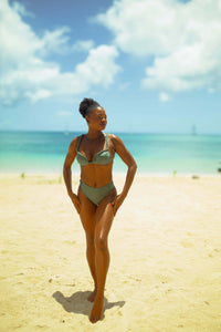 A woman in chic swimwear confidently showcases a flattering silhouette on a sandy beach against the backdrop of turquoise water. The partly cloudy sky enhances the bright and serene atmosphere as she wears her Claudine High-Waisted Bikini Bottom.