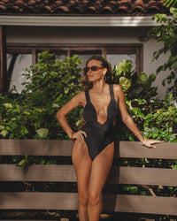 Woman in a black swimsuit standing on a wooden bench with a tiled roof and greenery in the background