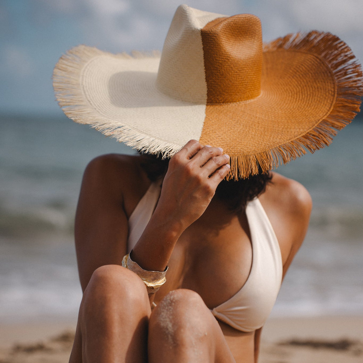 Woman sitting on a beach wearing a large two tone straw hat and white bikini.
