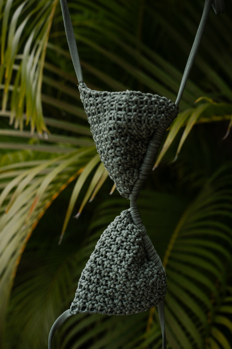 A close-up of the Rio Macrame Triangle Bikini Top, featuring textured green fabric with triangular cups and delicate straps, exuding handcrafted elegance against a backdrop of vibrant green palm leaves.