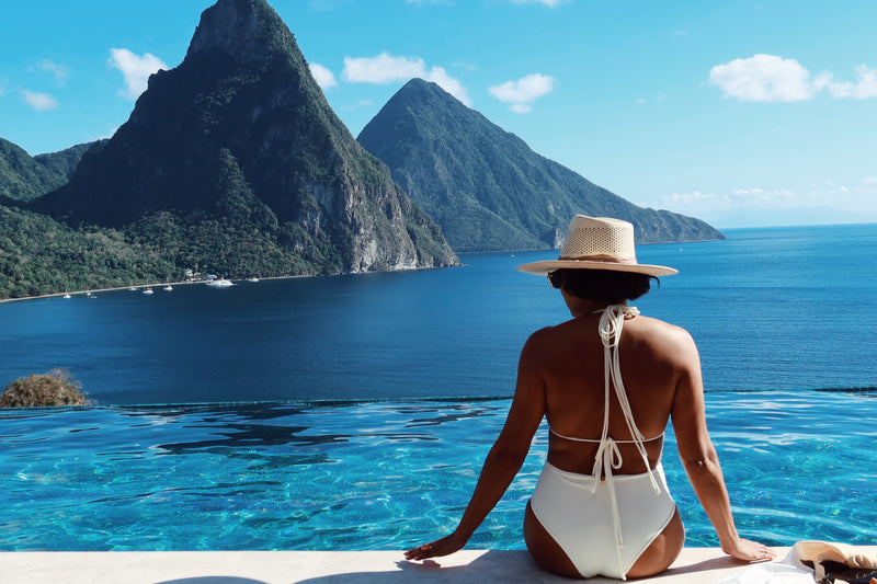 Wearing a stylish Dahlia Macramé One Piece Swimsuit and a straw hat, an individual relaxes beside an infinity pool with a breathtaking view of the sea and verdant mountains under a bright blue sky.