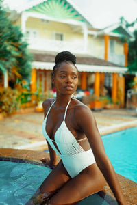 Wearing the Dahlia Macrame One Piece Swimsuit, a person sits by the pool's edge, gazing into the distance. The poolside is adorned with tropical plants, with a house featuring a decorative facade that showcases Colombian artistry in the background.