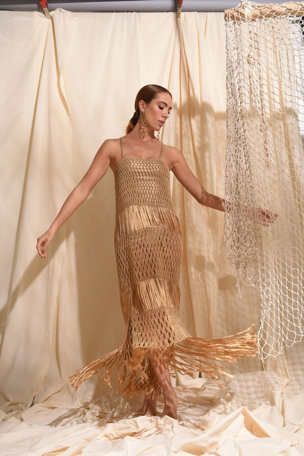 A woman stands gracefully in a room, adorned in the Dubai Macrame Dress. She poses with one hand outstretched and the other resting on a curtain, surrounded by luxurious gold yarns and soft beige fabric, embodying an elegant aura of handcrafted artistry.