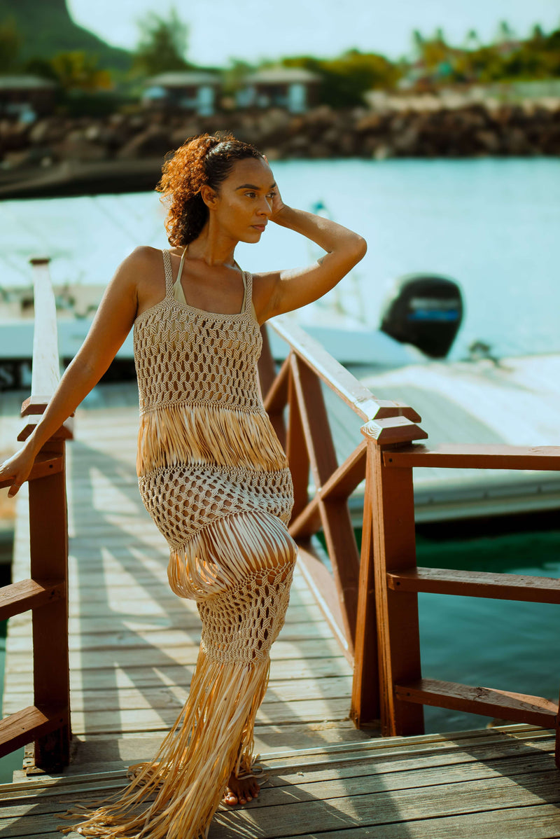 A person with curly hair is wearing the luxurious Dubai Macrame Dress, crafted from elegant gold yarns, as they stand on a wooden dock and lean against a railing. In the background, there is a body of water with moored boats and a rocky shoreline.