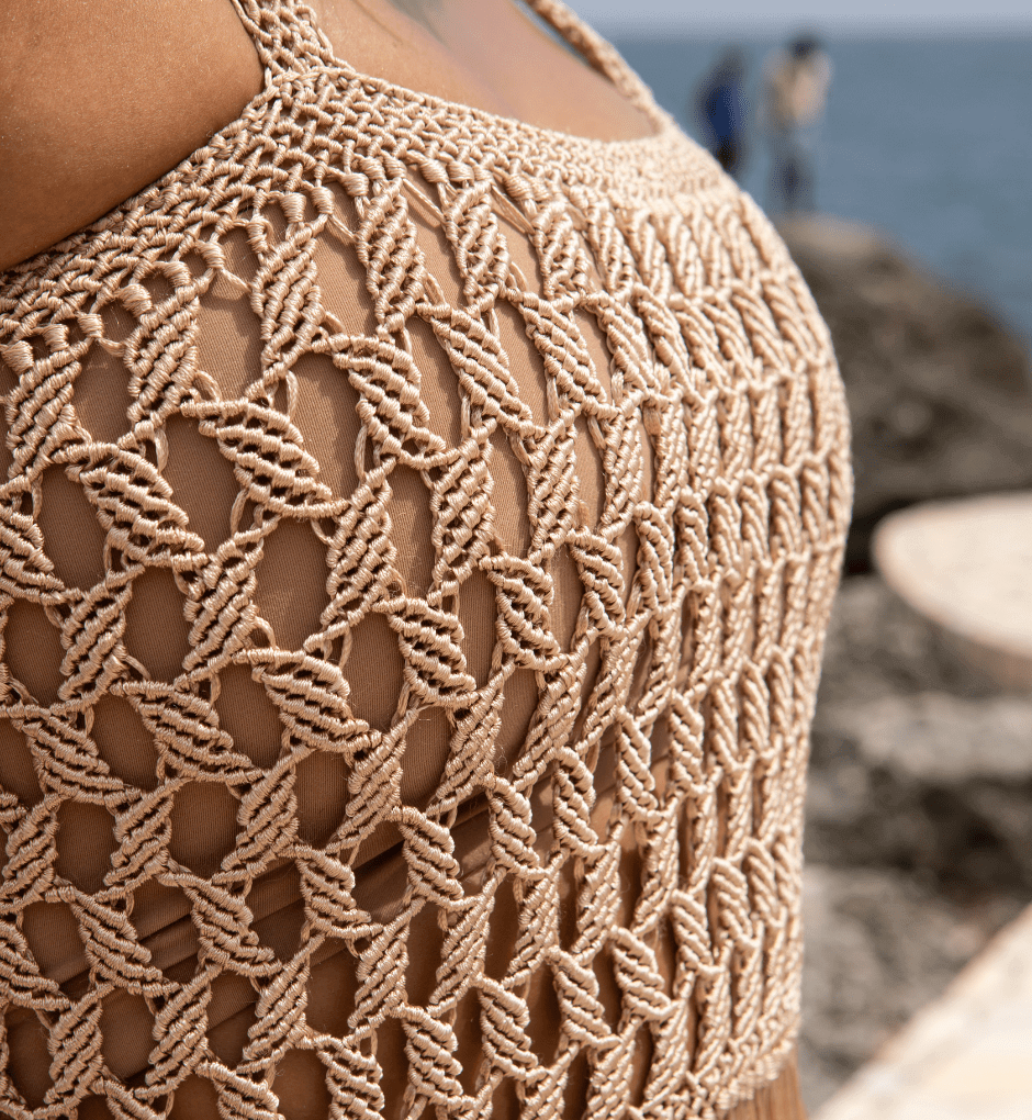 A close-up of an individual clad in a meticulously handcrafted Dubai Macrame Dress, featuring an elaborate woven design. In the background, blurred rocks and the ocean contribute to a coastal ambiance, with distant figures enhancing the depth of this scene.