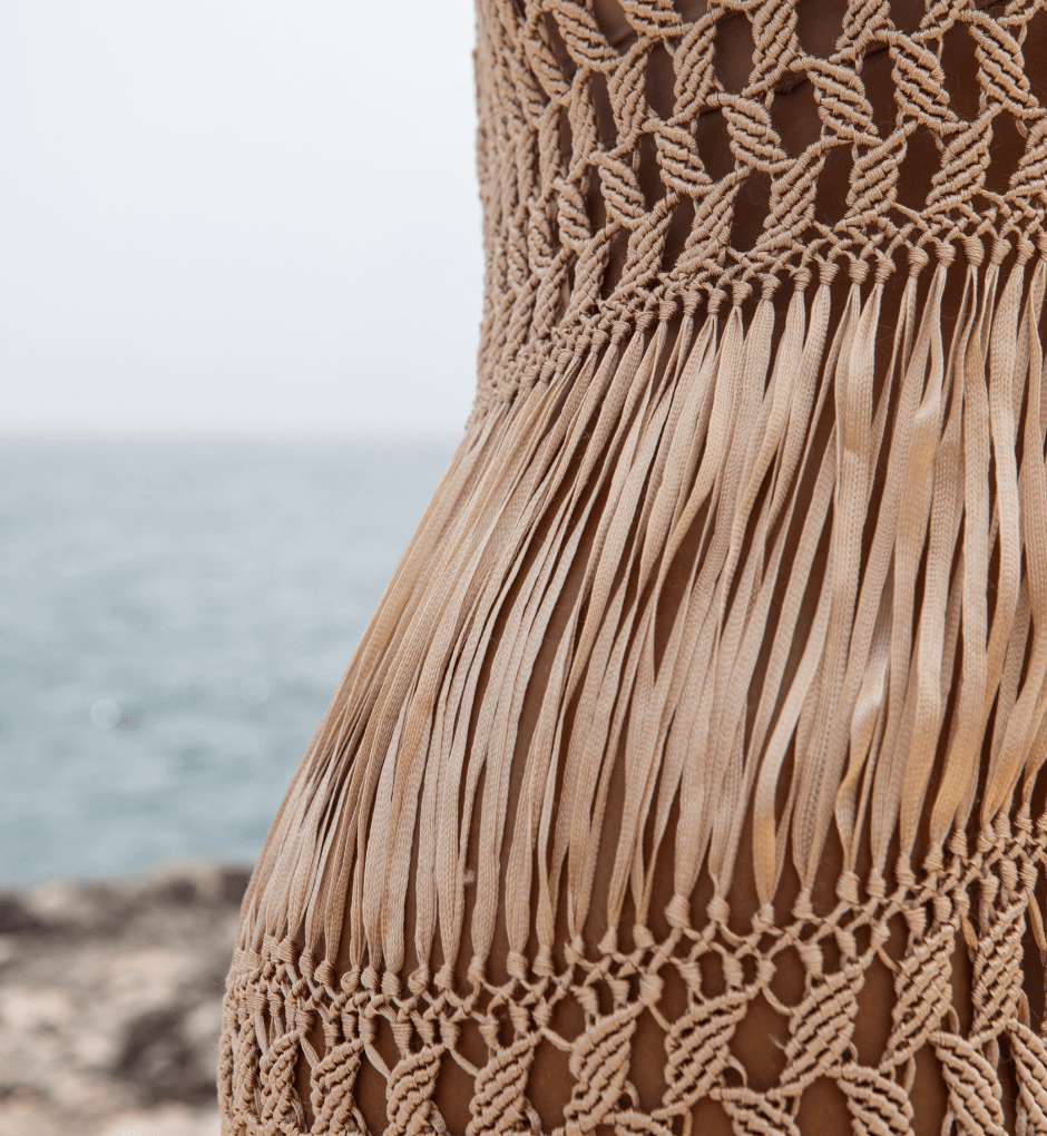 Close-up of the exquisite Dubai Macrame Dress, showcasing luxurious gold yarns and intricate fringe details against a blurred seascape background. The design masterfully combines open-knit and tightly woven sections, highlighting exceptional artisanal craftsmanship.