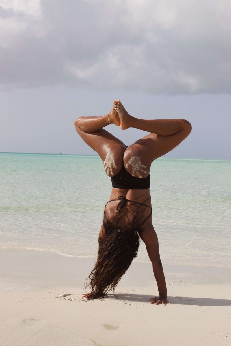 On a sandy beach with clear turquoise waters and cloudy skies in the background, someone elegantly performs a yoga handstand. They are wearing the Esa One Piece Swimsuit, crafted from luxurious Italian Carvico fabric, with sand visible on their legs.