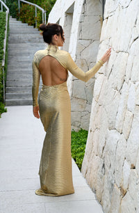 Wearing the Aurora Gold Maxi Dress, a woman radiates effortless glamour while posing outdoors. The dress's stunning backless design catches the light beautifully, accentuating its open details as she stands amidst a stone pathway surrounded by stairs and lush greenery.