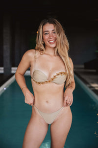 A woman with long hair smiles at the camera while standing at the edge of a pool. She is wearing a beige Fleur Handmade Bikini Top adorned with hand-beaded flowers, crafted in Colombia. Minimal jewelry complements her look, set against a dimly lit background with the pool’s water shimmering behind her.