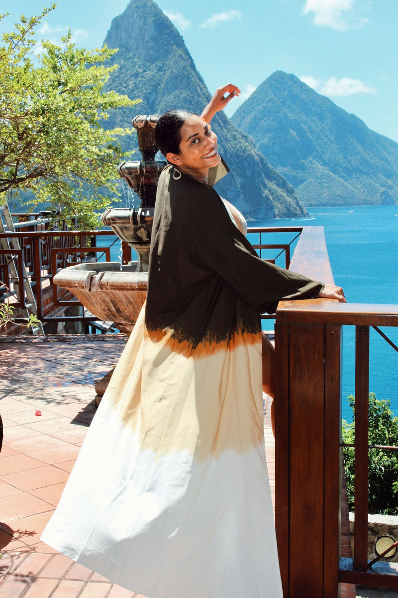 Wearing a Jakob Robe, a person stands smiling on a sunny terrace with wooden railings, overlooking a scenic view of two mountain peaks and the bright blue sea. Lush greenery surrounds the scene, perfectly complementing the vibrant summer style of their lightweight fabric attire.