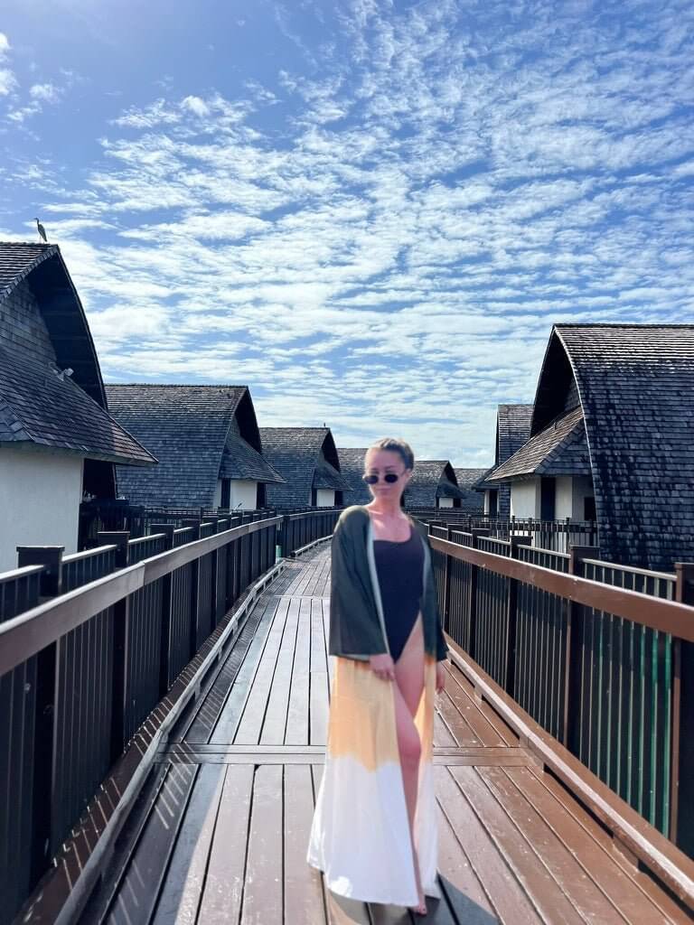 A person in sunglasses and the Jakob Robe stands on a wooden walkway lined with overwater bungalows. The lightweight fabric complements the bright sky with scattered clouds, capturing the essence of effortless summer style.