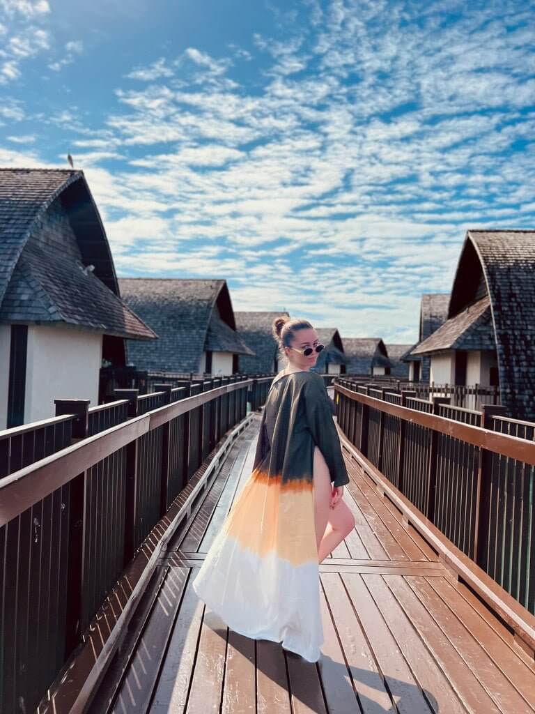 A person stands on a wooden walkway between overwater bungalows under a sky with scattered clouds, effortlessly capturing summer style. They wear sunglasses and the Jakob Robe, a long, flowing dress in shades of black, orange, and white made from lightweight fabric that dances gracefully in the breeze.