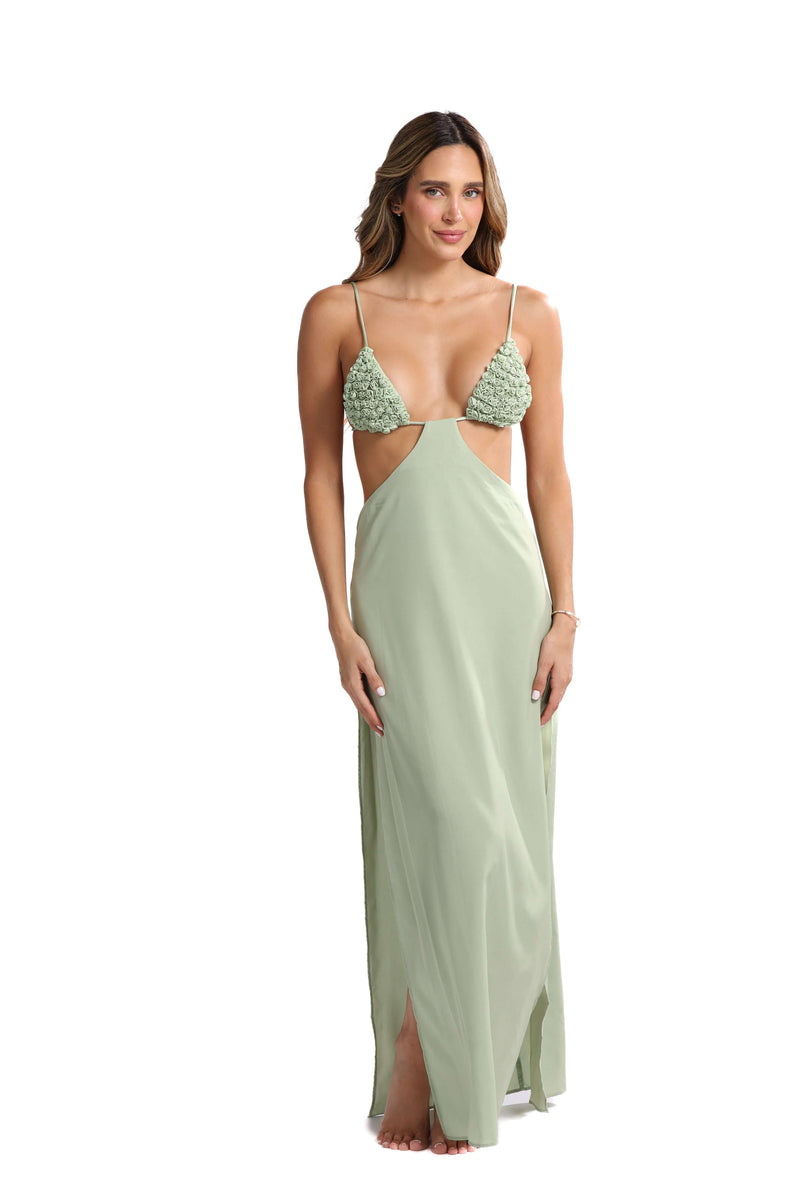 A woman poses against a white background wearing the Katt Clip On Maxi Skirt, designed in a light green hue with intricate, textured details at the top. Ideal for transitioning from beach to bar, this evening attire features thin straps, cut-out sides, and has a slightly sheer quality. Barefoot and gazing directly at the camera, she embodies effortless style.