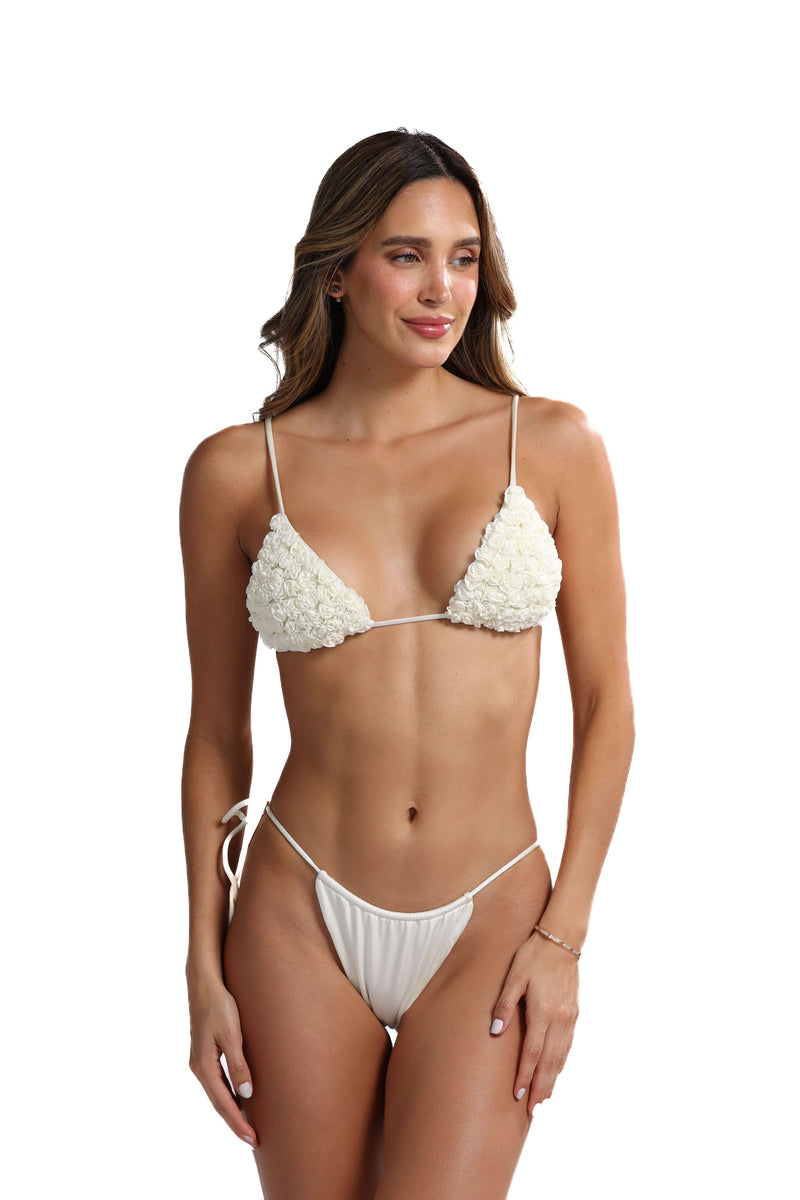 Wearing the Katt Triangle Bikini Top, which is adorned with a white floral pattern and string design featuring handcrafted mini rosettes, a person stands against a plain white background, looking to the side. This piece is an ideal addition to any swimwear collection.